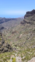 Steep green valley of Masca seen from Mirador de Masca, Tenerife. The serpentine road winds past village houses into a narrow ravine amid rugged cliffs, lush euphorbia and cacti, with the Atlantic on 