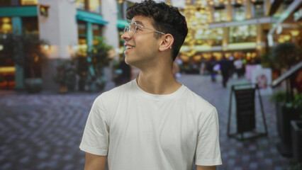 Fototapeta premium Man with curly hair and glasses turns his head to the side and smiles on a crowded city street, wearing a white t shirt; happiness.