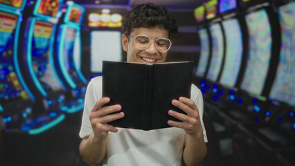 Man holding black hardcover book smiles broadly at slot machine reels in a neon lit building; contentment.