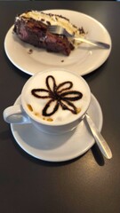 A white cup of cappuccino with milk foam and a chocolate flower design on a saucer with a spoon, with a slice of chocolate cake and a fork on a white plate in the background.