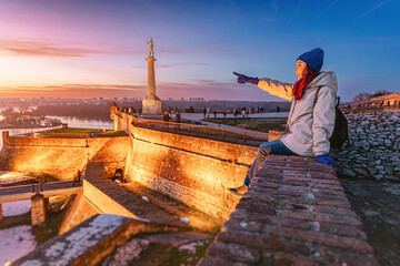 Young woman smiling and waving at sunset, observing Belgrade city skyline from Kalemegdan Fortress