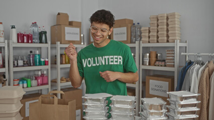 Young hispanic man smiles and points finger at stacked food containers in a building; compassion volunteerism.