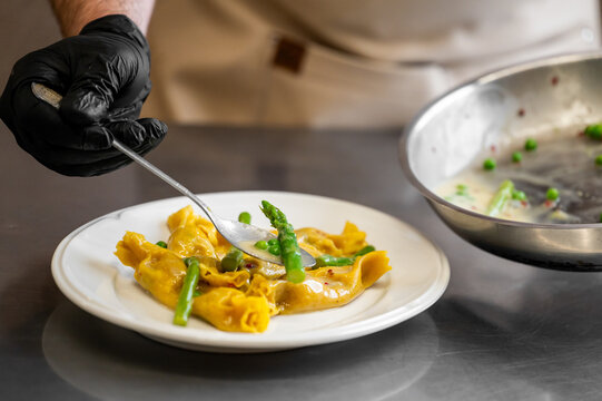 A chef wearing black gloves is plating a gourmet dish of fresh Italian agnolotti del plin pasta with asparagus and peas in a creamy butter sauce in a professional kitchen.