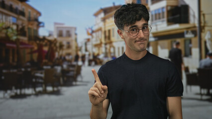 Young hispanic man points index finger upward on a sunlit city street beside outdoor cafe tables and blurred pedestrians; hesitation warning.