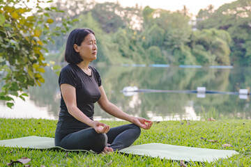 An Asian woman meditating beside a peaceful lake on a yoga mat, placed on fresh grass in a park...