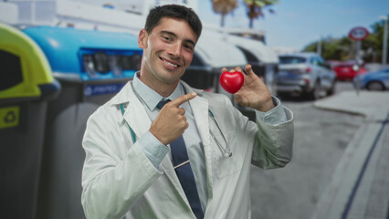 Man doctor in white coat with stethoscope points finger to small red heart held by other hand on...