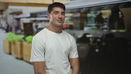 Man wearing white t-shirt smiling poses with arms resting on knee on restaurant terrace overlooking potted plants; relaxation.