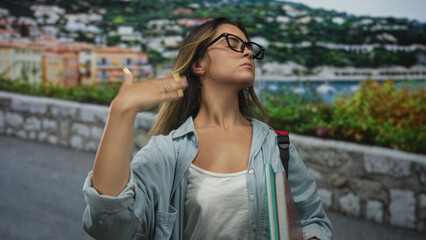 Young woman flipping hair while holding notebooks and backpack strap on a seaside street promenade, glasses on, looking up; confidence.