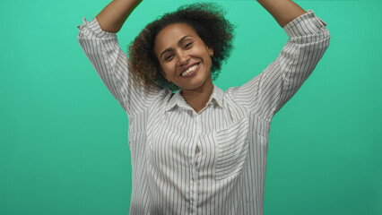 Woman smiling with arms raised showcasing bare forearms and a striped shirt in a teal studio; joy celebration.