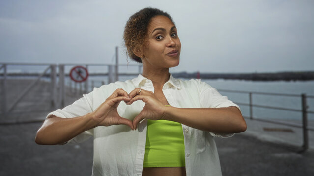 Woman forms heart gesture with hands on a street pier by the seaside, wearing lime crop top and white shirt while smiling at camera; love.