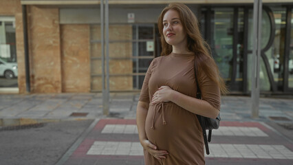 Fototapeta premium Pregnant woman in brown dress on city street crosswalk gently holds belly with one hand above and one hand below; anticipation.