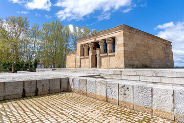 The Temple of Debod in Madrid, an ancient Egyptian monument, features two weathered stone pylons and the main structure, set on a stone plaza against a background of trees and a cloudy blue sky