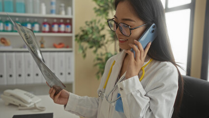 Young chinese woman doctor in uniform with a stethoscope talking on the phone while examining an x-ray in a clinical setting, showcasing a professional healthcare environment.