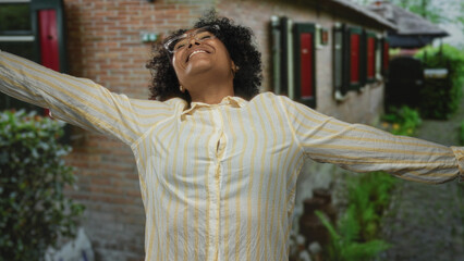 Laughing woman wearing yellow striped shirt spreads arms outside sunlit building entrance; pure joy.