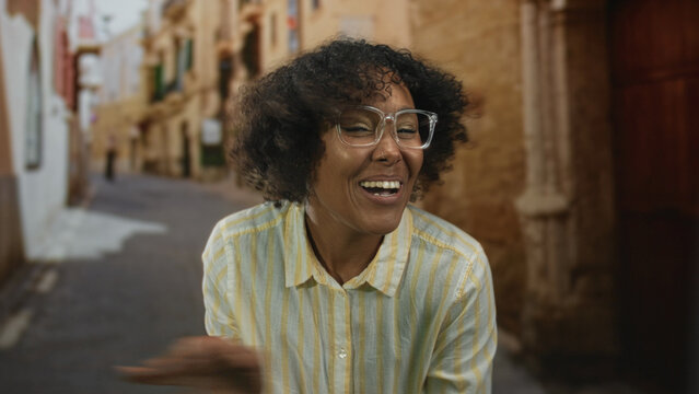 Woman with glasses and curly hair smiles on old stone street with narrow cobblestone path and historic buildings; joy. - Powered by Adobe