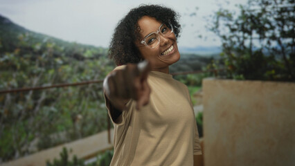 Woman wearing beige shirt smiles and shapes heart with fingers in outdoor building setting; love warmth joy connection.