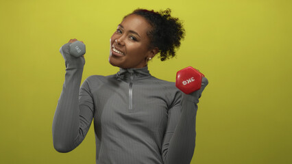Athletic woman lifts a pair of red and gray dumbbells and smiles in a green studio space; confidence.