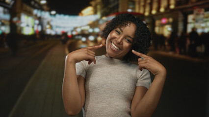 Woman smiling and pointing finger to mouth on urban street lit by storefront lights at night; happiness.