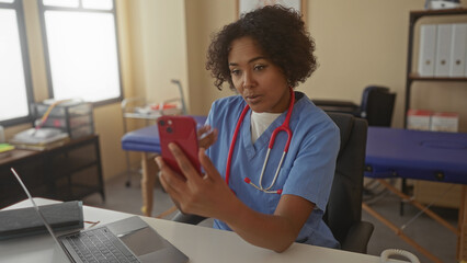 Woman doctor in blue scrubs holds red smartphone and touches chest while seated at desk with laptop in clinic; empathy trust.