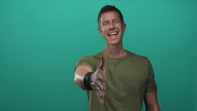 Man extending right hand for handshake and smiling in a teal studio with olive t shirt and bracelets; friendly welcome.