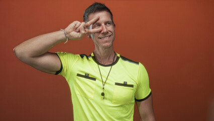 Man referee displaying peace sign with hand over eye in orange studio setting; playful sporty confidence.