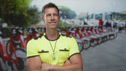 Man with arms crossed, whistle on a lanyard and neon shirt at bike rental parking on a city street; confidence urban service.