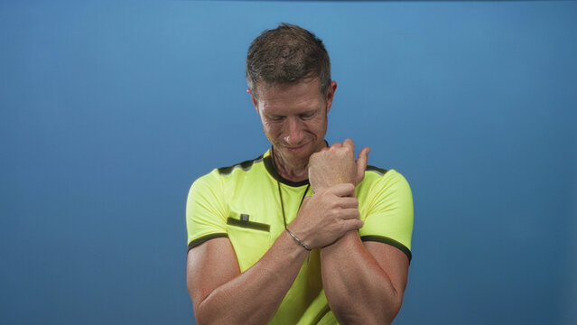 Man referee clutching wrist and forearm with both hands in studio set with blue wall; determination.