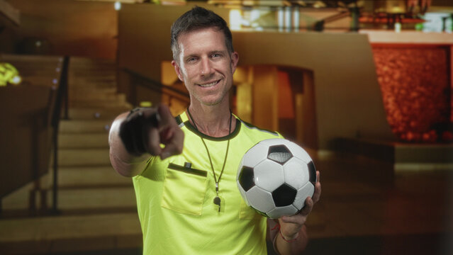Young man wearing yellow shirt holding soccer ball and points finger toward lens in building lobby by staircase; confidence team encouragement. - Powered by Adobe
