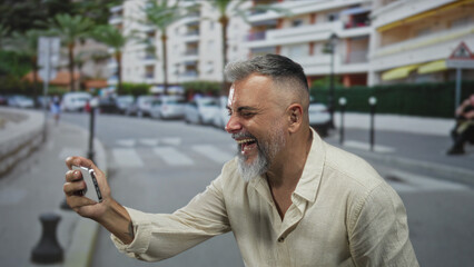 Man holding smartphone with both hands, laughing and looking at the screen while standing on a busy city street crosswalk near parked cars;joy connection.