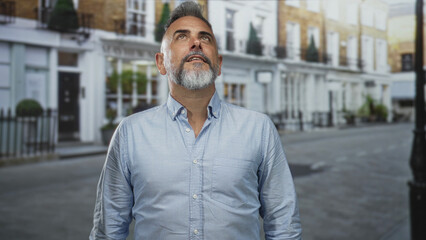 Fototapeta premium Man smiling with eyes closed, gray beard visible, standing on a quiet city street in front of townhouse storefronts; quiet contentment.
