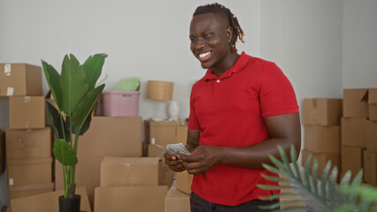 Man counts cash with hands in building surrounded by unpacked moving boxes and houseplants; moving day joy.