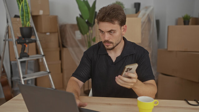 Man holding smartphone and typing on laptop with hands amid packed cardboard boxes and ladder in building; concentration moving home. - Powered by Adobe