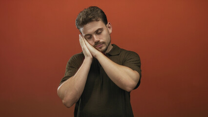 Young hispanic man with hands pressed to cheek sleeping gesture in studio with orange backdrop wearing dark polo shirt; calm rest.
