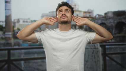 Fototapeta premium Man with hands cupping ears at building ruins on an outdoor balcony, looking upward and tensing shoulders; frustration focus.