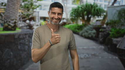 Man smiling and showing thumbs up with his bare hand in a building courtyard with greenery and pathway visible; optimism everyday life.
