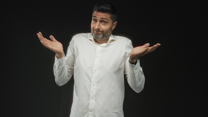 Man shrugging with open hands and raised shoulders in a black studio, casual white shirt and trimmed beard, questioning expression; uncertainty ambivalence.