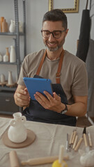 Man taps blue tablet at pottery table in studio wearing apron and glasses while seated among tools and ceramics; calm contentment.