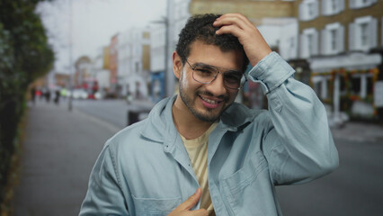 Man with glasses wearing a denim jacket smiles as he touches hair with a bare hand on a busy city...