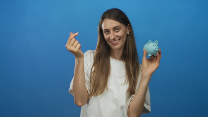 Woman holding blue piggybank making finger heart gesture with hands and smiling in studio; saving...
