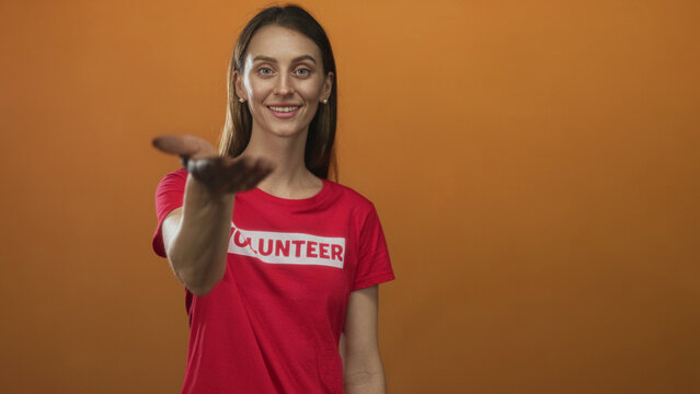 Young caucasian woman volunteer wearing red volunteer t shirt holds out her hand palm up toward camera in orange studio; community support hopeful.