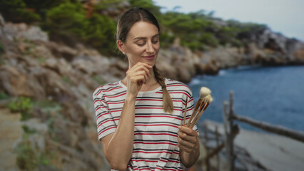 Woman holding paintbrushes and touching chin at seaside near rocky coast in striped shirt and braid; contemplation.