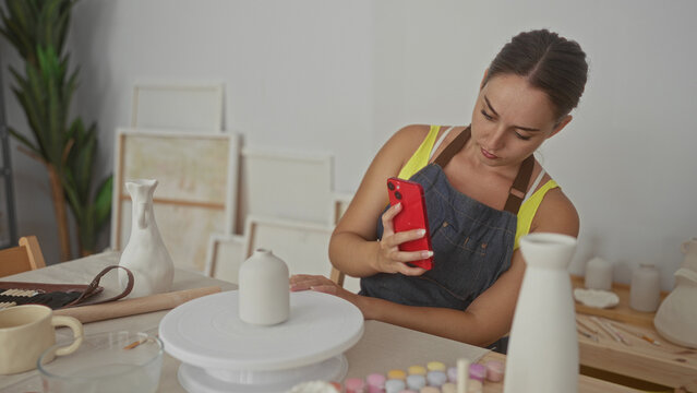 Woman holding red smartphone and touching clay on pottery wheel in studio; concentration creativity craft. - Powered by Adobe