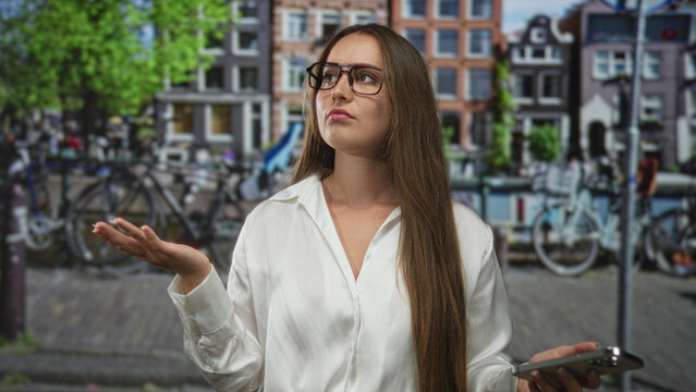 Woman wearing glasses and white shirt holding smartphone with palm up gesture on street in amsterdam; frustration.