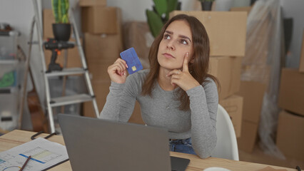 Woman holding creditcard with hand on chin while working at laptop in building; budget planning thoughtful.