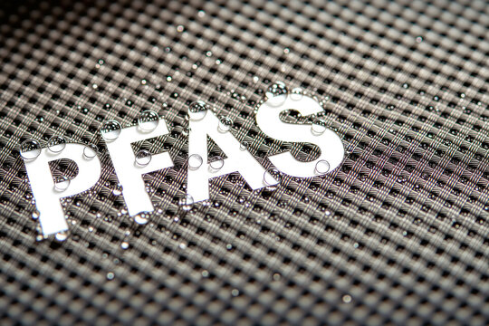 The acronym PFAS is displayed in white text on a black and white checkered water-resistant fabric covered with small, spherical water droplets.