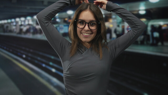 Woman smiling with hands on head, face visible, wearing glasses and grey shirt on a crowded train station platform indoors; casual joy.