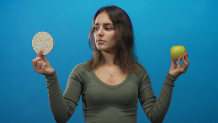 Woman holding green apple and rice cake in blue studio, arms extended to each side while comparing...