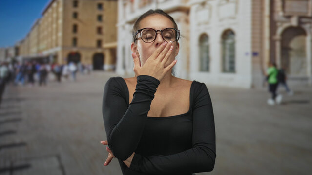 Young woman in black top and glasses with fist at chin and hand covering mouth on a city street near a historic building; tired contemplation. - Powered by Adobe