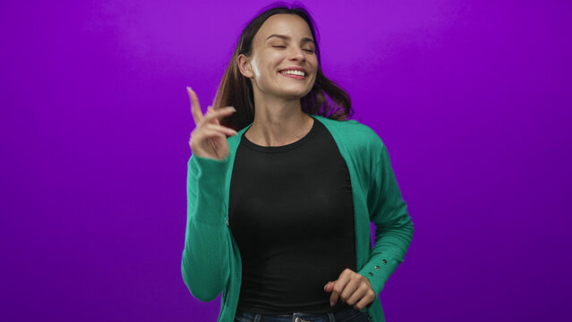 Woman pointing fingers while dancing in a purple studio with solid violet wall and closed eyes enjoying motion; joyful playful groove.