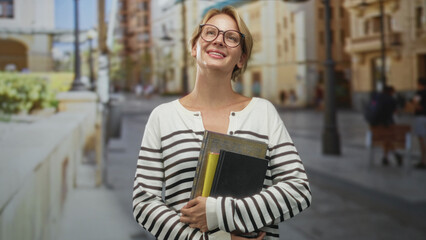 Fototapeta premium Blond woman with glasses holds books and a black notebook to her chest smiling with head tilt on a sunny city street lined with buildings and lampposts; joy study confidence.